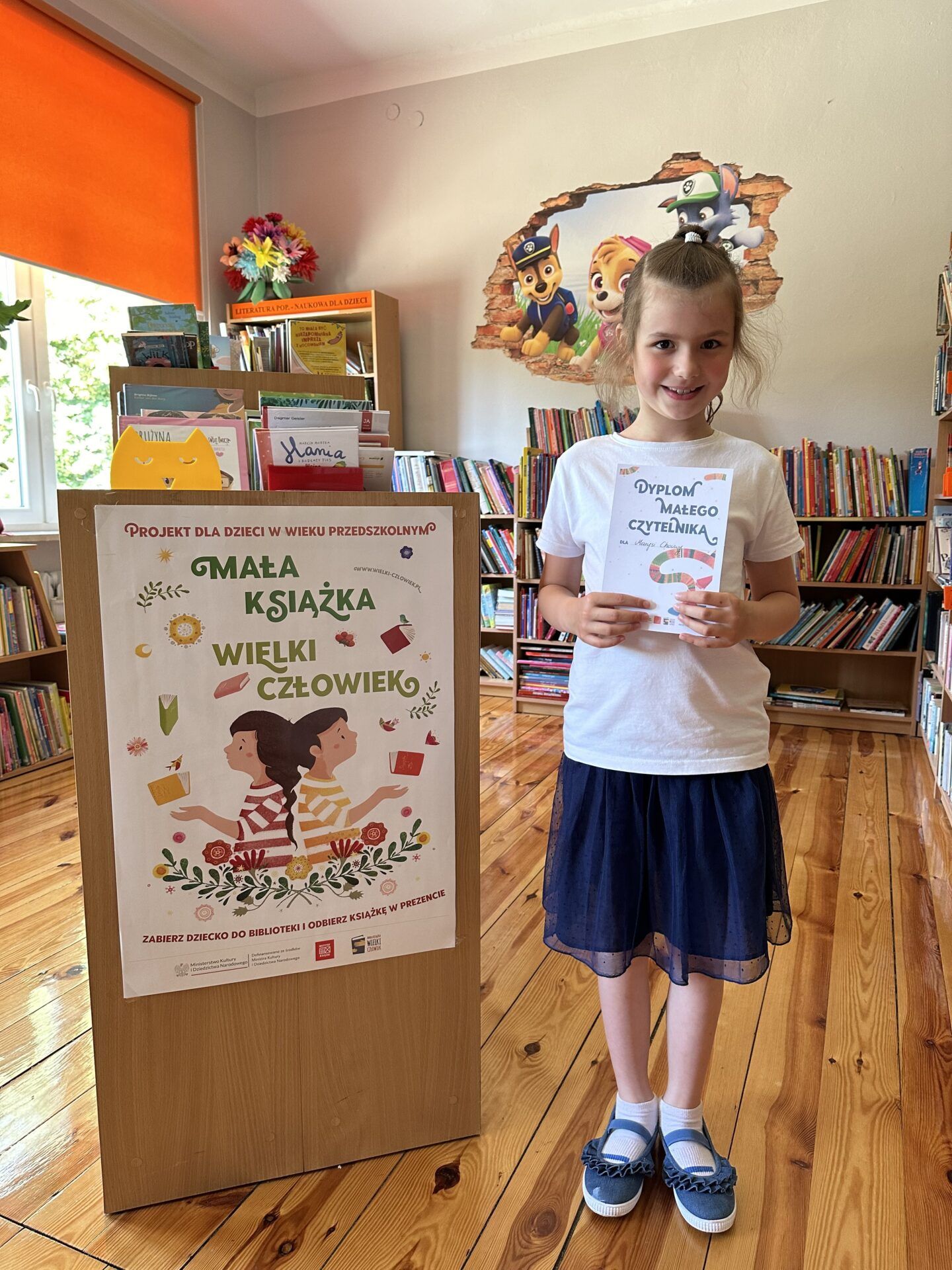 Dziewczynka z Dyplomem Małego Czytelnika na tle półek z książkami w wypożyczalni dla dzieci.