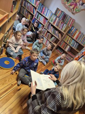 przedszkolaki między regałami w bibliotece słuchający książki czytanej przez bibliotekarkę
