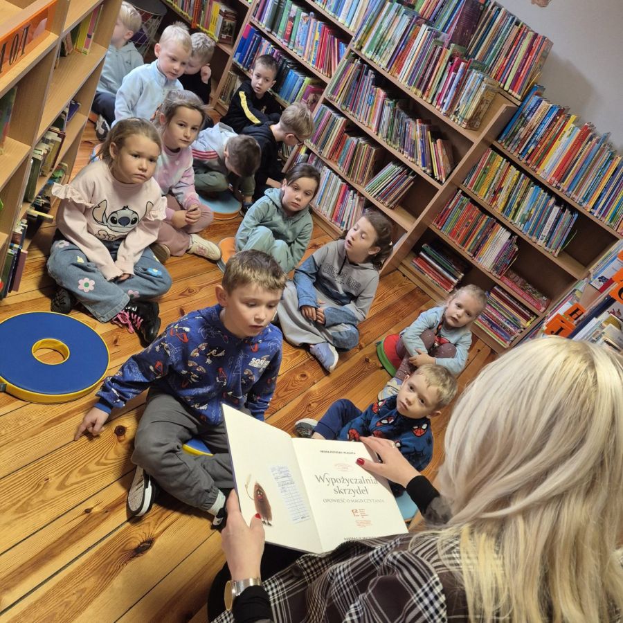 przedszkolaki między regałami w bibliotece słuchający książki czytanej przez bibliotekarkę