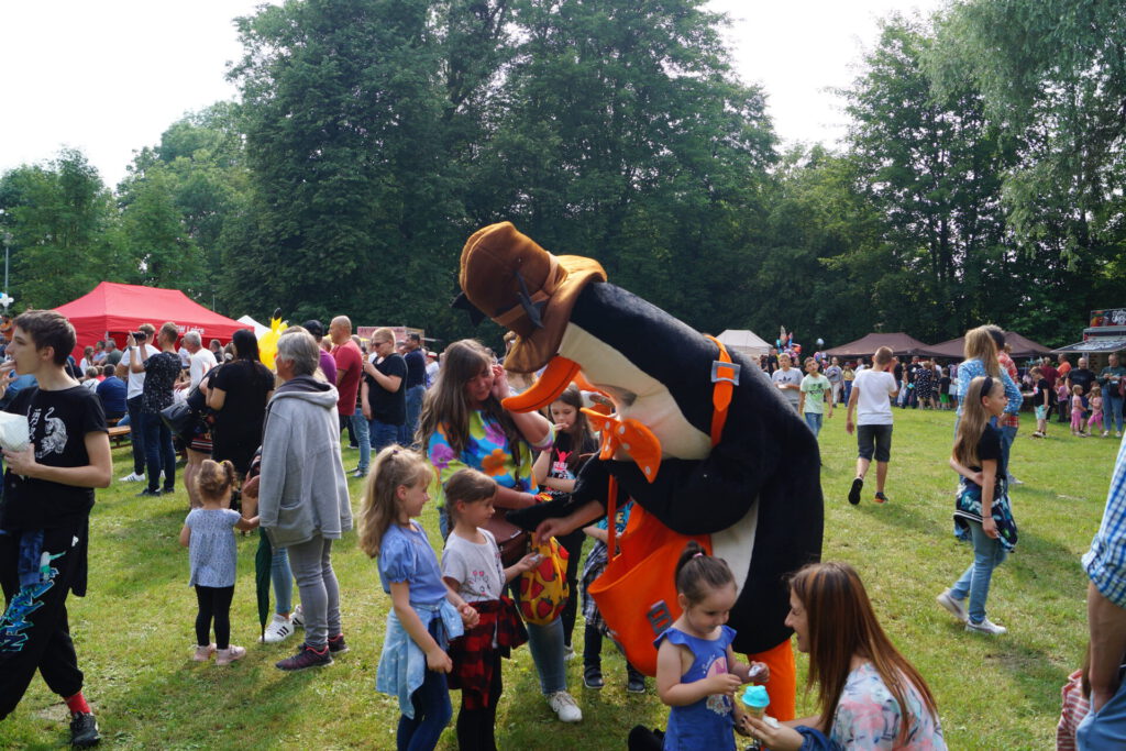 Maskotka "Pingwin" rozdająca cukierki dzieciom na tle uczestników festynu rodzinnego.