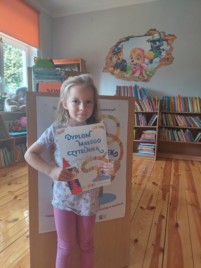 Lena (uczestniczka projektu "Mała książka - wielki człowiek") trzymająca dyplom na tle wypożyczalni książek dla dzieci.