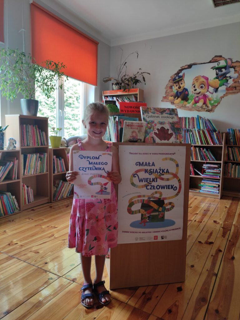 Blanka (uczestniczka projektu "Mała książka - wielki człowiek") trzymająca dyplom na tle wypożyczalni książek dla dzieci.