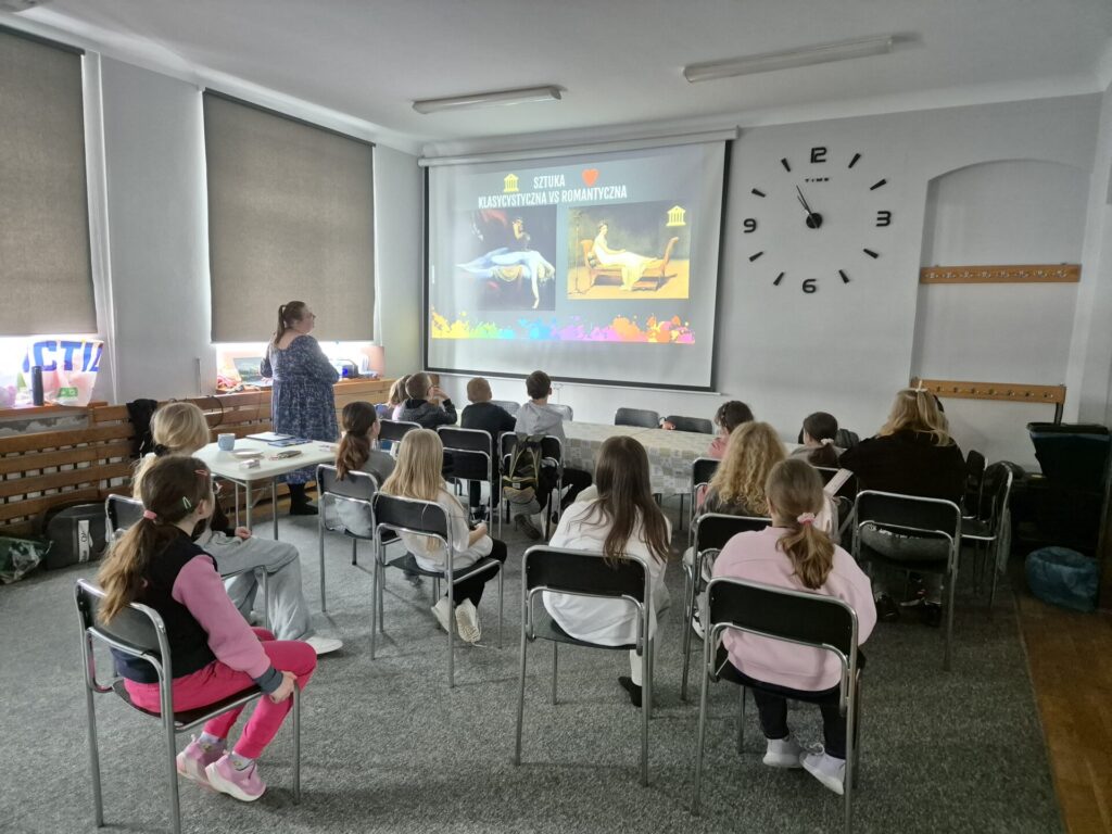 14 dzieci siedzących na krzesełkach i oglądających prezentację wyświetloną przez opiekunką na ekranie 