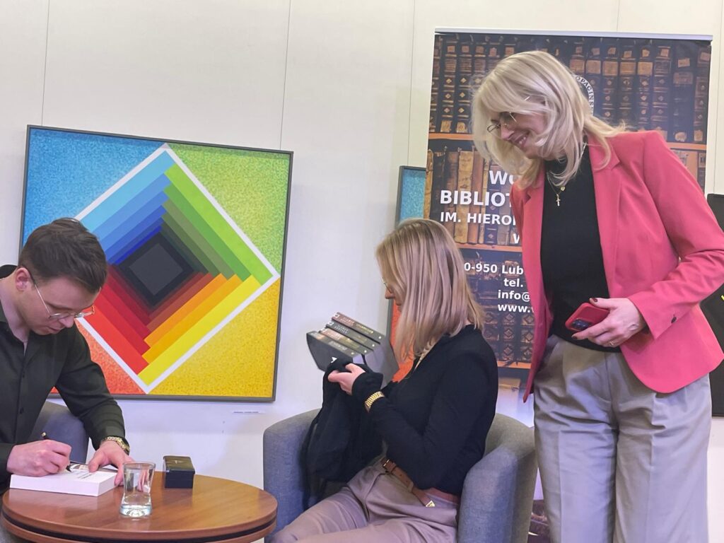 Mężczyzna podpisujący książkę i dwie kobiety podczas spotkania autorskiego.