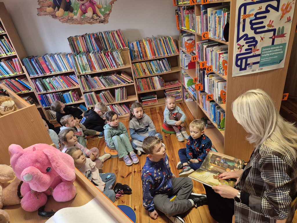 przedszkolaki między regałami w bibliotece słuchający książki czytanej przez bibliotekarkę