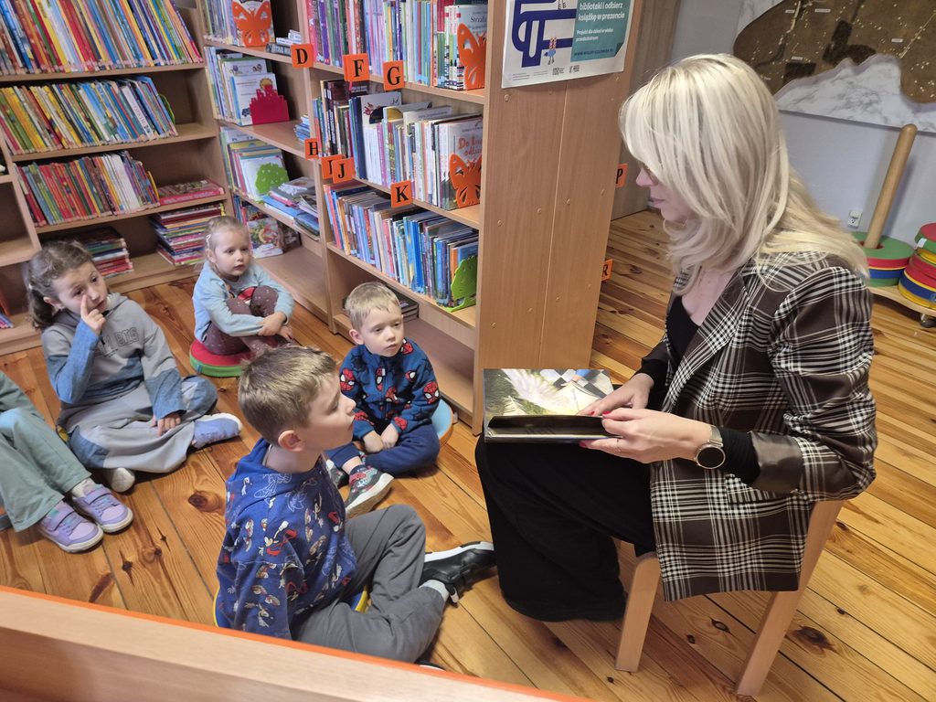 przedszkolaki między regałami w bibliotece słuchający książki czytanej przez bibliotekarkę