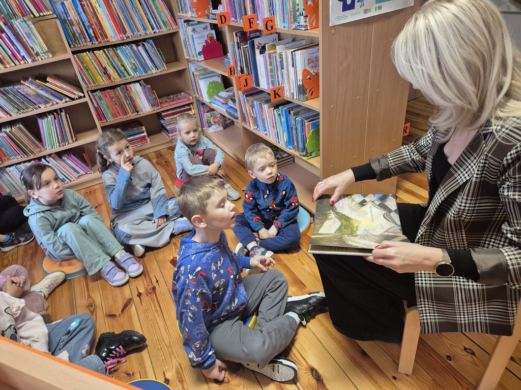 przedszkolaki między regałami w bibliotece słuchający książki czytanej przez bibliotekarkę