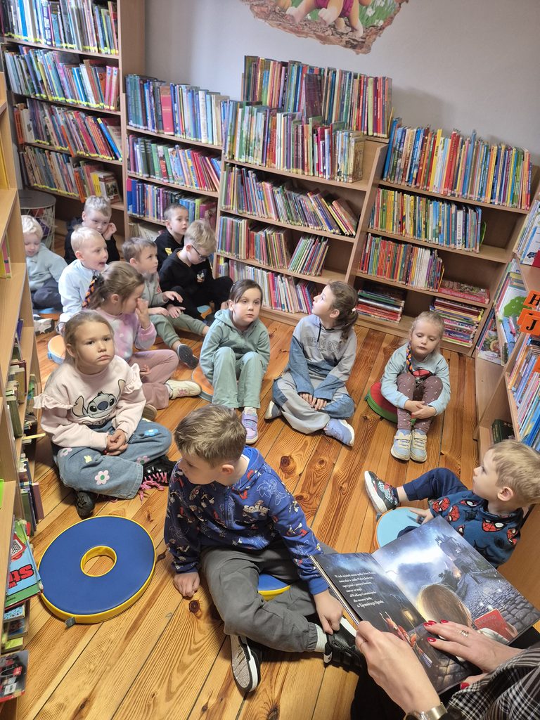 przedszkolaki między regałami w bibliotece słuchający książki czytanej przez bibliotekarkę